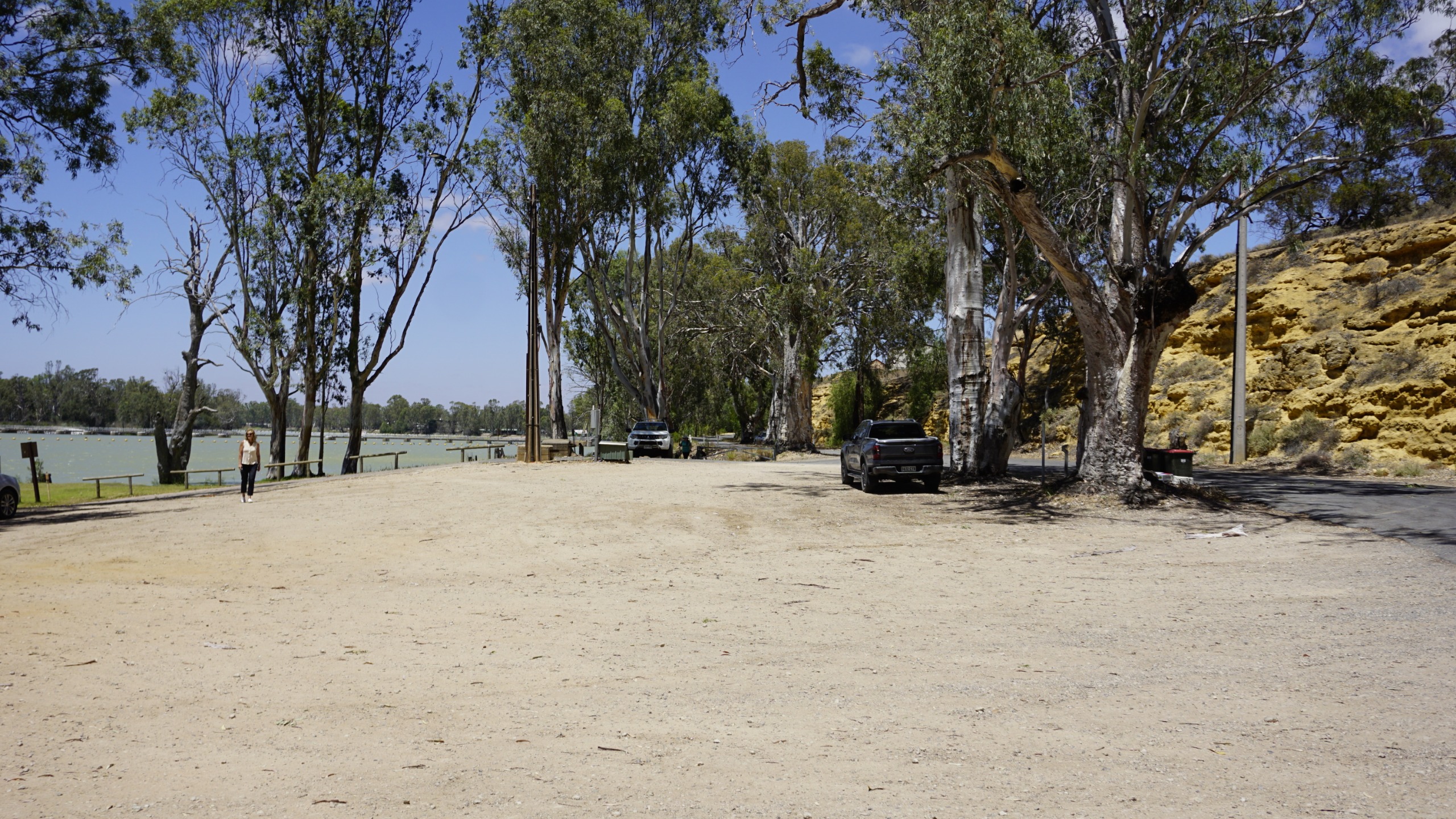 Before: A wide outdoor riverside space with a large, open dirt area in the foreground. Tall gum trees line the edge of the river, casting light shade. On the left, a calm, pale-green river with floating buoys stretches into the distance. A single person is walkin