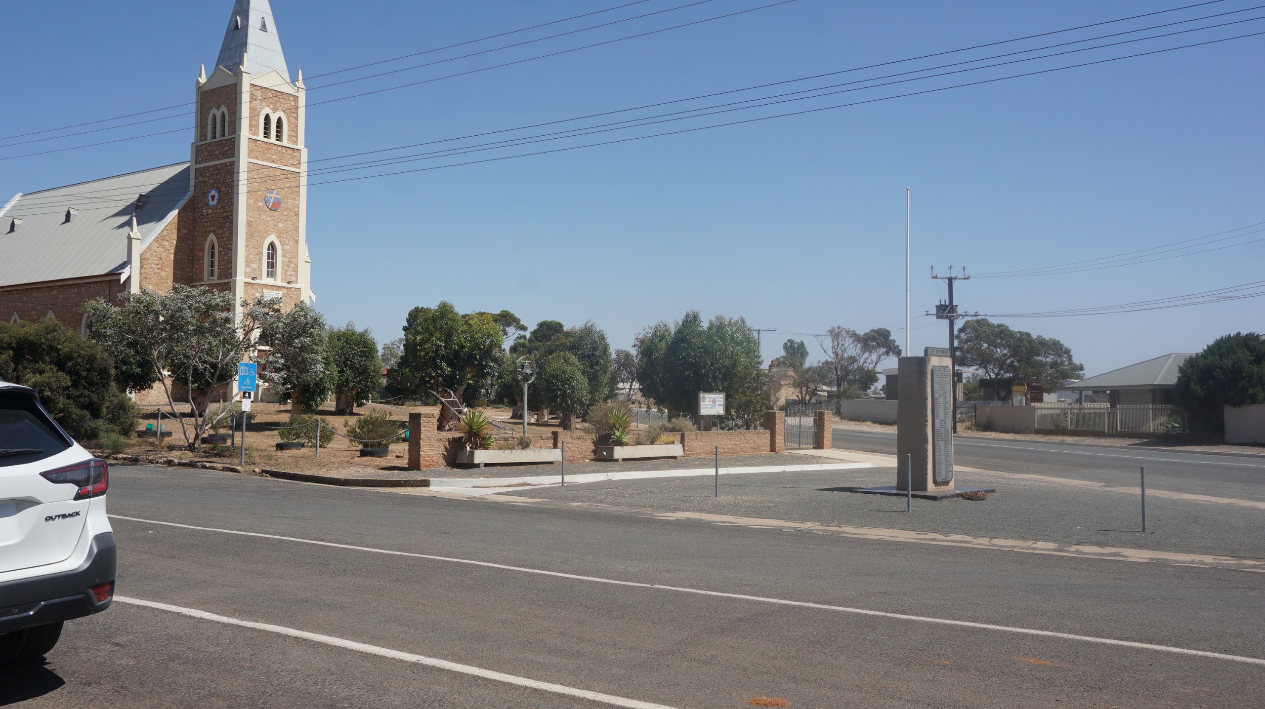 Before: A quiet rural town intersection under a clear blue sky, featuring a historic stone church building on the left with a tall steeple and arched windows. The church sits on slightly raised ground with dry landscaping, low shrubs, and small trees surrounding 
